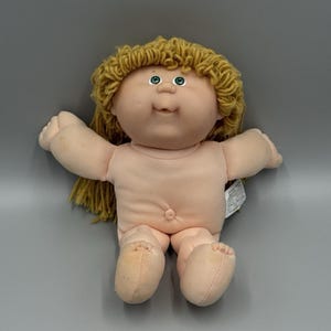 May include: A Cabbage Patch Kids doll with a soft fabric body, light skin tone, and yarn hair in a golden-brown shade. The doll features green eyes, a small nose, and a smiling mouth. It has outstretched arms and legs, and a white tag is visible.