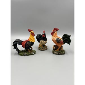 May include: Three rooster figurines standing on textured green bases. The left rooster has yellow, red, and black feathers. The center rooster is black and white. The right rooster features orange, red, and dark green plumage with a red comb.