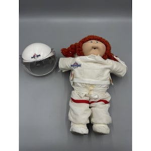 Può includere: Una bambola con capelli di lana rossa in trecce, che indossa una tuta da astronauta bianca con strisce rosse e un logo ricamato, accompagnata da un casco separato in plastica bianca e trasparente con lo stesso logo da astronauta.