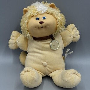 May include: A plush cat doll with light yellow fabric, blue eyes, and a brown collar featuring a round gold-toned metal tag engraved with the name CRIS. The doll has yarn-like hair in a lighter shade of yellow and a soft, rounded body shape.