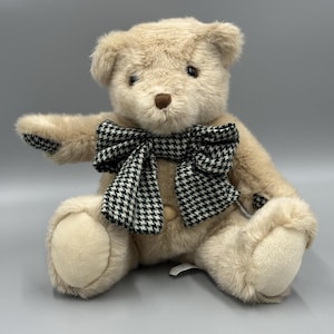 May include: A plush teddy bear with light brown fur and a black and white houndstooth bow tie. The bear is sitting with its arms outstretched, and has a button on its chest.