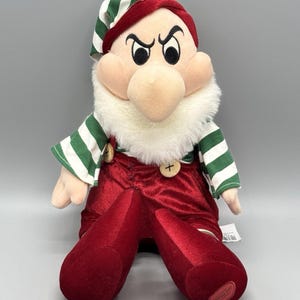 May include: A plush doll of a character with a large nose, thick white beard, and furrowed eyebrows. The doll wears a red velvet-like jumpsuit with two light-colored buttons and a green and white striped shirt and matching pointed hat.