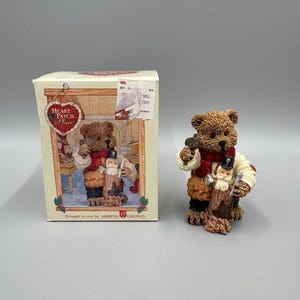 Könnte beinhalten: Eine Heart Patch Place-Figur eines braunen Bären mit rotem Schal, der eine kleine Figur hält. Die Figur steht neben der Originalverpackung mit dem Heart Patch Place-Logo und dem Schriftzug "Brought to you by AMERICAN GREETINGS."