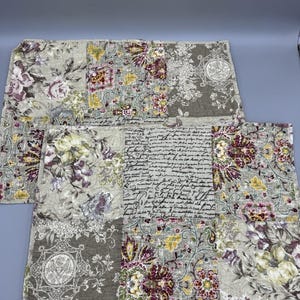 May include: Two rectangular placemats with a patchwork design. The placemats feature floral patterns in shades of purple, yellow, and green on a beige background. Some patches include script-like text and decorative medallions.