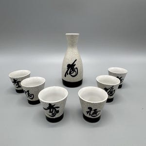 Puede incluir: Un juego de sake con una botella de cerámica texturizada de color blanquecino y siete tazas a juego. Cada pieza tiene una base negra y un carácter japonés negro. El juego está diseñado para servir sake.