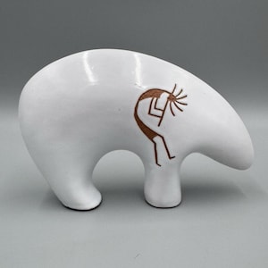 May include: A smooth, white, bear-shaped ceramic figurine featuring a brown, etched Kokopelli figure on its side. The bear is depicted in a standing position with its head bowed downward, showcasing a clean, glossy finish and simple, rounded lines.