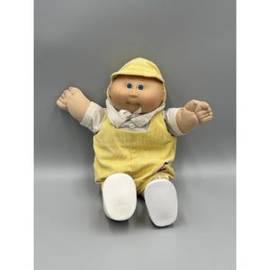 Peut inclure: Une poupée en tissu avec un visage rond, des yeux bleus et des cheveux en laine, portant une casquette en velours côtelé jaune et une salopette assortie sur une chemise blanche à col, avec des chaussures blanches et un petit motif brodé.