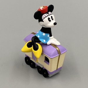 May include: A Minnie Mouse figurine sits atop a purple and cream-colored train car. Minnie wears a blue polka-dot dress, yellow shoes, and a red hat. The train car has gold trim and black wheels.