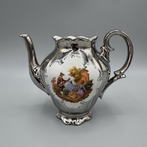 May include: A silver-toned ceramic teapot featuring a central white oval panel with a colorful illustration of two figures in a landscape. The teapot has a scalloped rim, a curved handle, and a spout, with ornate silver detailing surrounding the scene.