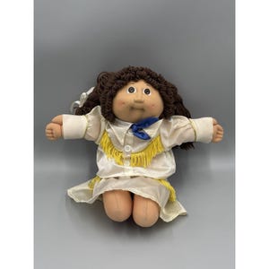 Op de afbeelding: Een Cabbage Patch Kids pop met donkerbruin wollen haar, bruine ogen en kuiltjes in de wangen. De pop draagt een crèmekleurig shirt met lange mouwen en een rok met gele franjes, witte knopen en een kleine blauwe halsdoek.