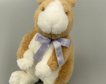 May include: A plush stuffed animal in the shape of a rabbit with soft tan and white fur. The rabbit is in a seated position and features a light purple satin ribbon tied in a bow around its neck. The fur is textured and short, with white patches on the face, chest, and feet.
