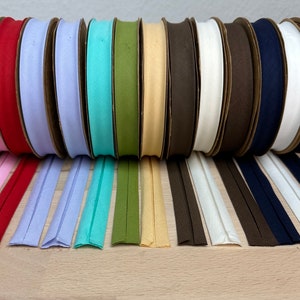 May include: A collection of 12 spools of cotton bias tape in various colors, including purple, pink, red, light blue, turquoise, green, beige, brown, white, and navy blue. The tape is rolled up on cardboard spools and laid out in a row on a wooden surface.