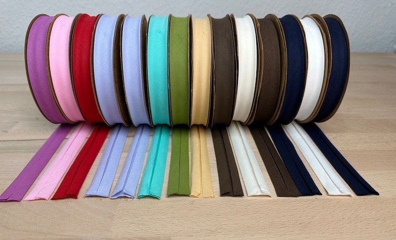 May include: A collection of 12 spools of cotton bias tape in various colors, including purple, pink, red, light blue, turquoise, green, beige, brown, white, and navy blue. The tape is rolled up on cardboard spools and laid out in a row on a wooden surface.