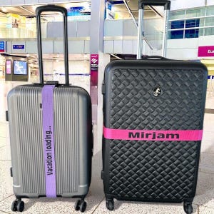 Può includere: Due valigie con cinturini per bagagli. La valigia a sinistra è grigia con un cinturino viola che dice "vacation loading..." La valigia a destra è nera con un cinturino rosa che dice "Mirjam".