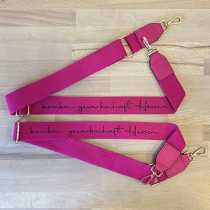 Könnte beinhalten: Zwei rosa verstellbare Riemen mit schwarzem Text, der "komba gewerkschaft hessen" lautet.