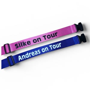 Könnte beinhalten: Zwei Stoff-Armbänder mit schwarzen Kunststoffverschlüssen. Eines ist rosa mit "Silke on Tour" in Schwarz bedruckt. Das andere ist blau mit "Andreas on Tour" in Weiß bedruckt. Beide sind auf weißem Hintergrund abgewinkelt.
