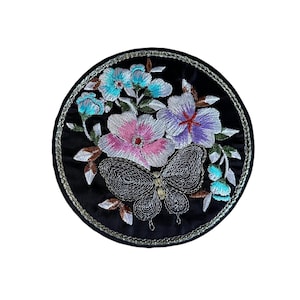Peut inclure: Un patch circulaire noir avec des fleurs brodées et un papillon. Les fleurs sont roses, violettes et bleues avec des accents argentés. Le papillon est argenté avec des accents noirs.