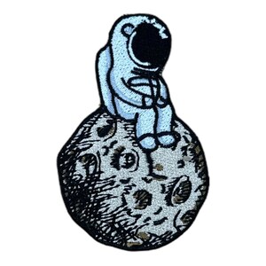 Könnte beinhalten: Ein gestickter Aufnäher mit einem blauen Astronauten, der auf einem grauen Mond mit Kratern sitzt. Der Astronaut trägt einen weißen Raumanzug mit einem schwarzen Helm.