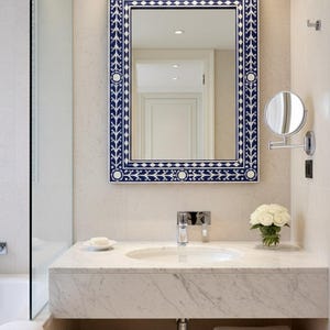 Puede incluir: Un mueble de baño con encimera de mármol y un gran espejo cuadrado. El espejo tiene un borde decorativo azul y blanco. Un pequeño lavabo, grifo y un jarrón con flores blancas están sobre la encimera. Hay toallas blancas apiladas debajo.