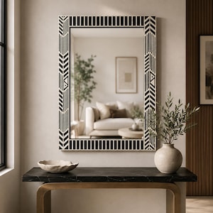 Puede incluir: Un espejo de pared rectangular con un marco de patrón geométrico en blanco y negro que consiste en rayas verticales y flechas en chevrón. El espejo refleja el interior de una sala de estar con un sofá, una planta en maceta y un cuadro.