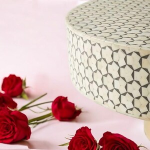 May include: A white round table with a black geometric pattern. The table is made of bone inlay and has a gold base. The table is surrounded by red roses.
