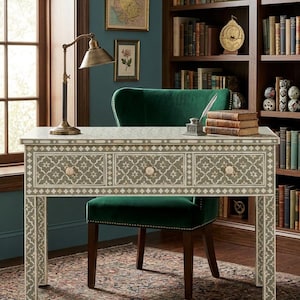 Puede incluir: Un escritorio decorativo con un patrón geométrico gris y blanco, tres cajones y pomos de color rosa claro. Una silla de terciopelo verde está detrás del escritorio. Una lámpara de escritorio de latón y libros están sobre el escritorio. La habitación tiene una pared azul oscuro y una alfombra estampada.