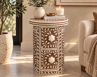 May include: A cylindrical side table featuring a brown and white floral vine inlay pattern. The table has two stacked drawers, each equipped with a white, flower-shaped knob. The top surface holds a white vase, a small bowl, and a book titled The KINFOLK Home.