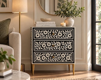May include: A three-drawer chest featuring a black finish with white floral inlay patterns and gold-toned hardware. The chest stands on four tapered gold legs and is topped with two books titled The Kinfolk Home and At Home, a bowl, and a vase.