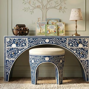 Può includere: Una scrivania blu navy e bianca con motivo floreale e uno sgabello abbinato. La scrivania ha cassetti e un arco curvo per lo sgabello. Un'arca giocattolo in legno, libri e una lampada con un design a luna e stella sono sulla scrivania. La stanza ha un tema di asilo nido.
