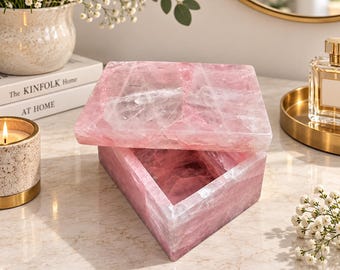 May include: A square box crafted from polished rose quartz, featuring a removable lid. The stone displays natural translucent pink hues with white crystalline veining throughout the surface. The box has a smooth, glossy finish and a geometric shape.