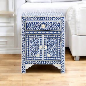 May include: A rectangular bedside table featuring a vibrant blue and white floral inlay pattern. The piece includes one top drawer and two lower cabinet doors, each equipped with small, round, off-white flower-shaped knobs for opening.