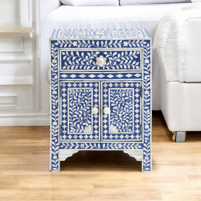 May include: A rectangular bedside table featuring a vibrant blue and white floral inlay pattern. The piece includes one top drawer and two lower cabinet doors, each equipped with small, round, off-white flower-shaped knobs for opening.