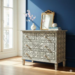 Puede incluir: Cómoda decorativa con un patrón de sol en blanco y negro. Cuenta con cuatro cajones y un espejo con marco dorado. Un jarrón blanco con flores rosas se encuentra encima. La habitación tiene una pared azul y una ventana.