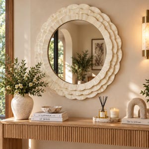 Op de afbeelding: Een ronde wandspiegel met een gelaagd, geschulpt frame in een lichte, crèmekleurige, getextureerde afwerking. De spiegel reflecteert een binnenruimte met een plant en het frame bestaat uit drie concentrische, overlappende cirkelvormige lagen.