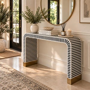 May include: A rectangular console table featuring a black and white striped pattern across its entire frame, with solid gold-toned metal bases at the bottom of each leg. The table surface holds a white vase, a bowl, two books, and a candle.