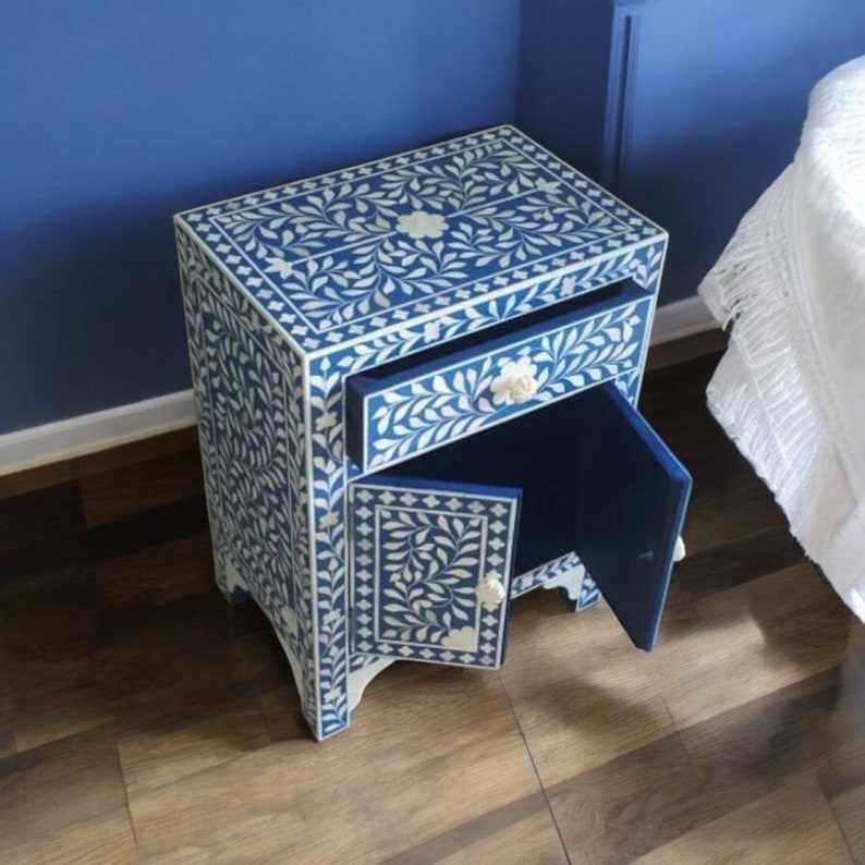 May include: A rectangular bedside table featuring a deep blue finish with intricate white floral and leaf inlay patterns. The piece includes one pull-out drawer and one cabinet door, both equipped with small, round, off-white floral-shaped knobs.