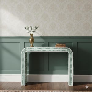 May include: A white and green console table with a curved design, featuring a geometric pattern. The table is set against a backdrop of green paneling and floral wallpaper. A gold vase with greenery and books are on top.
