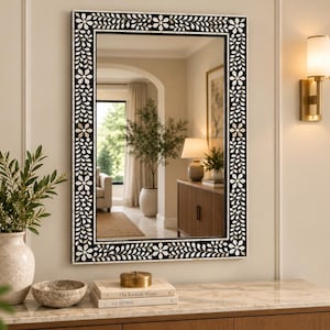 May include: A rectangular wall mirror featuring a black frame with an intricate white floral and vine inlay pattern. The mirror reflects a room interior, and the frame displays a repeating design of small flowers and leaves along all four sides.
