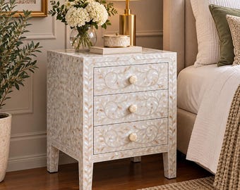 May include: A three-drawer bedside table featuring an intricate cream and white floral inlay pattern across the front and sides. The piece includes three round, textured knobs and stands on four straight legs with a matching inlaid finish.