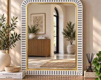 May include: A rectangular wall mirror with rounded corners, featuring a frame with a repeating pattern of vertical grey and white stripes and a thin gold inner border. The mirror reflects a room with a wooden cabinet, a potted plant, and a rug.