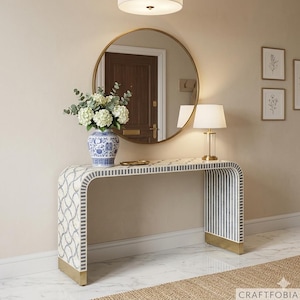 Puede incluir: Una consola decorativa con un diseño curvo. La mesa presenta una parte superior con estampado blanco y gris, con un patrón de rayas en los laterales. Tiene detalles metálicos dorados en las patas. Un espejo redondo cuelga encima de la mesa y una lámpara está encima.