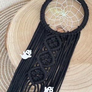Könnte beinhalten: Schwarzer Makramee-Traumfänger mit weißem Netzdesign und drei weißen geisterförmigen Anhängern. Der Traumfänger hat einen kreisförmigen Rahmen und lange, schwarze Fransen. Das Design umfasst drei kreisförmige Makramee-Knoten.