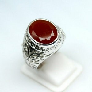 May include: A silver ring with an oval, faceted, dark red gemstone. The ring has an ornate, engraved design around the band. The gemstone is set in a bezel setting, and the ring is displayed on a white surface.