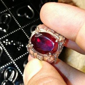 Può includere: Un anello d'argento con una gemma ovale di colore rosso intenso. L'anello ha un'incastonatura ornata e strutturata. La gemma è il punto focale, catturando la luce e mostrando un colore ricco e scuro. L'anello è tenuto dalle dita di una persona.