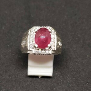 May include: A silver ring with a large, oval ruby gemstone. The ruby is a deep red hue, encircled by smaller, clear gemstones. The ring has a detailed, textured band and is displayed on a white stand.