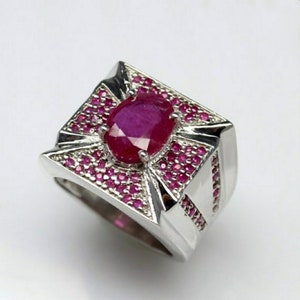 May include: A silver ring with a large, oval-shaped ruby gemstone at the center. The ring is surrounded by smaller, round rubies and has a geometric design. The ring is set against a white background.