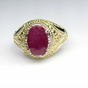 May include: A silver and gold ring featuring a large, oval-cut ruby gemstone. The ruby is a deep red color and is set in a decorative bezel. The ring has a detailed, textured design around the gemstone.