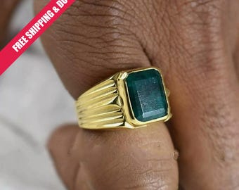 May include: A gold ring featuring a rectangular, emerald-cut green gemstone set in a polished gold band with a ribbed, textured design along the sides. The ring has a substantial, structured appearance with a prominent, deep green center stone.