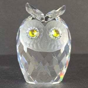 Puede incluir: Figura de búho de cristal transparente con cuerpo y alas facetadas. El búho tiene ojos de cristal amarillo y transparente y una cara esmerilada. La figura es un artículo decorativo.