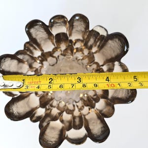 May include: A brown glass vase with a unique, flower-like design. The vase is measured with a tape measure, showing a diameter of approximately 10 inches.